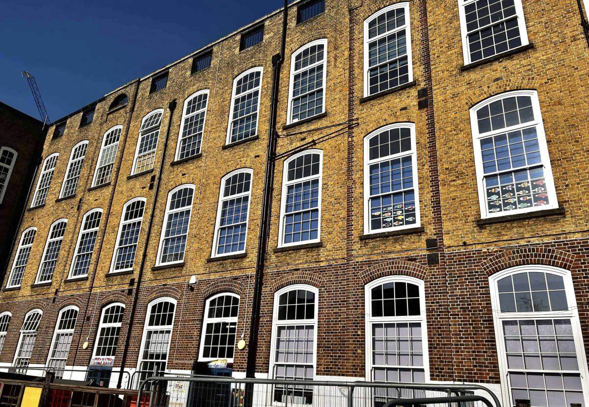 A tall brick building with large arched windows under a clear blue sky, featuring a metal fence in the foreground.