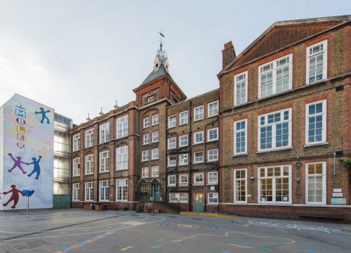 A traditional brick school building stands beside a modern annex with vibrant climbing figures on it, under a clear sky.