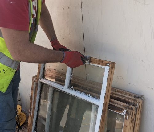 A person in a hi-vis vest and gloves repairs a window frame against a wall, with other frames nearby.