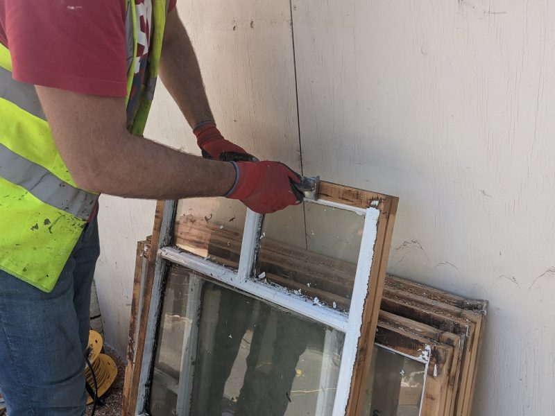 A person in a hi-vis vest and gloves repairs a window frame against a wall, with other frames nearby.