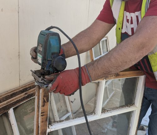 A person wearing red gloves uses a power tool on a window frame outside a beige building.