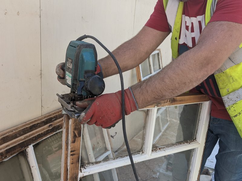 A person wearing red gloves uses a power tool on a window frame outside a beige building.