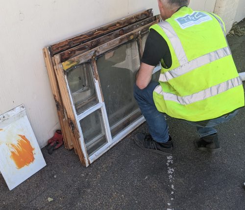 A person in a high-visibility vest is squatting beside a pile of old wooden windows, inspecting them, against an exterior wall.