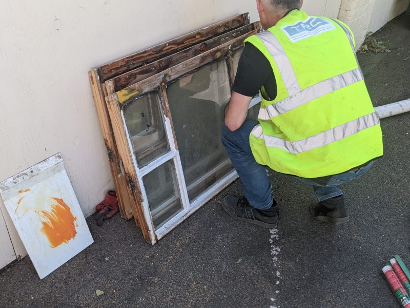 A person in a high-visibility vest is squatting beside a pile of old wooden windows, inspecting them, against an exterior wall.