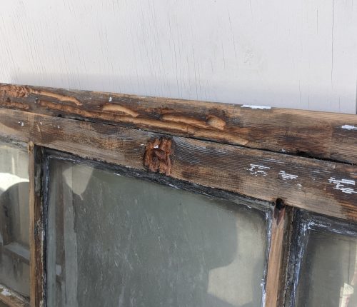 A weathered wooden window frame with peeling paint, showing signs of decay and damage, set against a white wall.