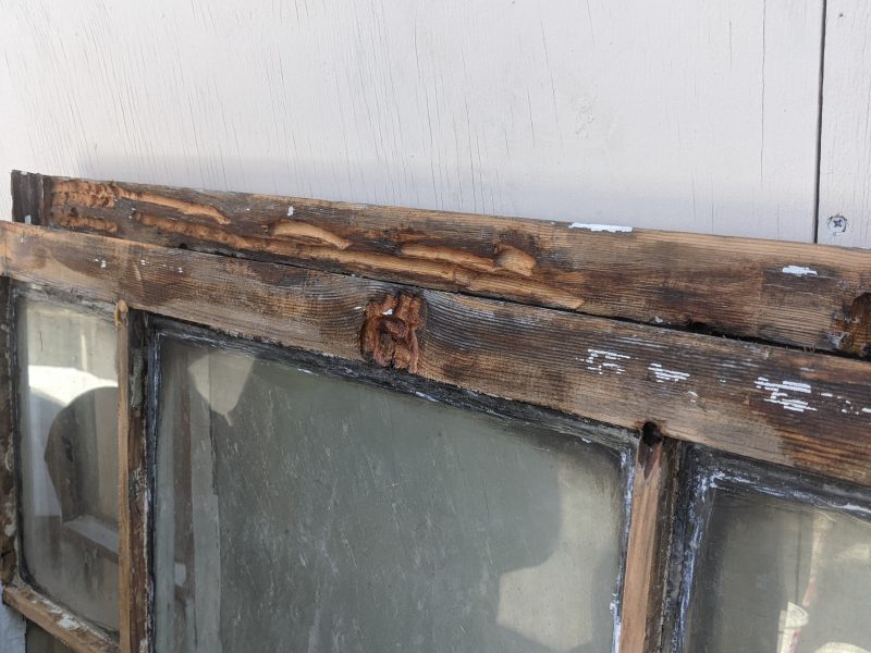 A weathered wooden window frame with peeling paint, showing signs of decay and damage, set against a white wall.
