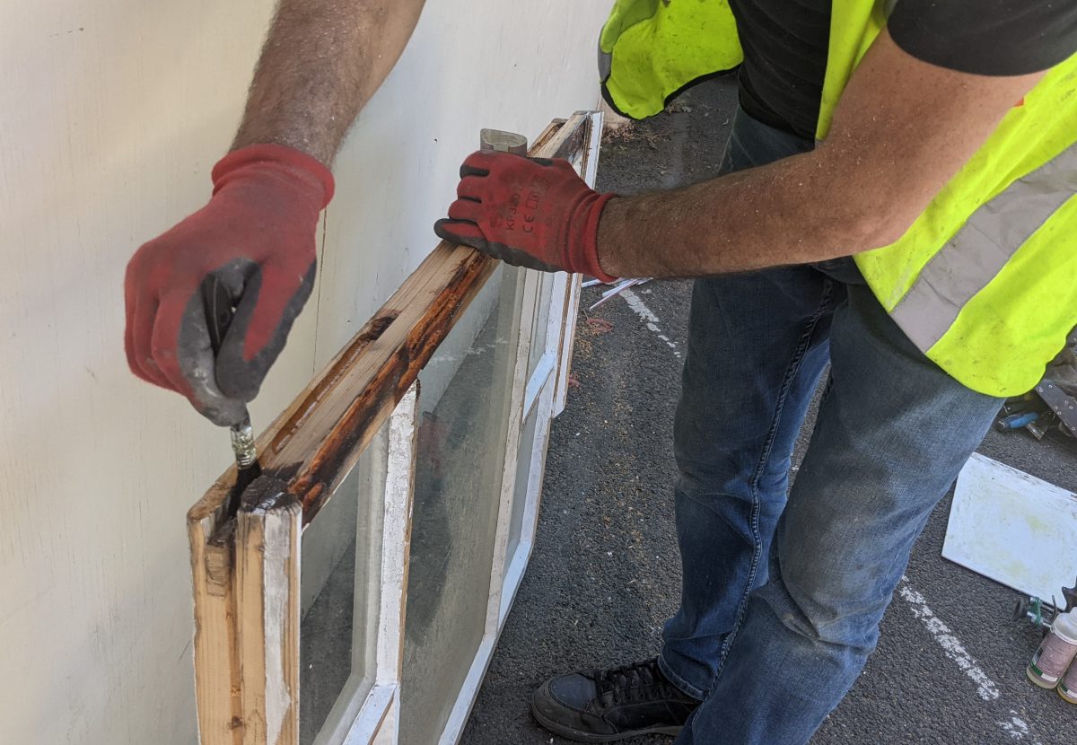 A person in safety gear uses pliers on a window frame, repairing it, with construction debris and a wall in the background.