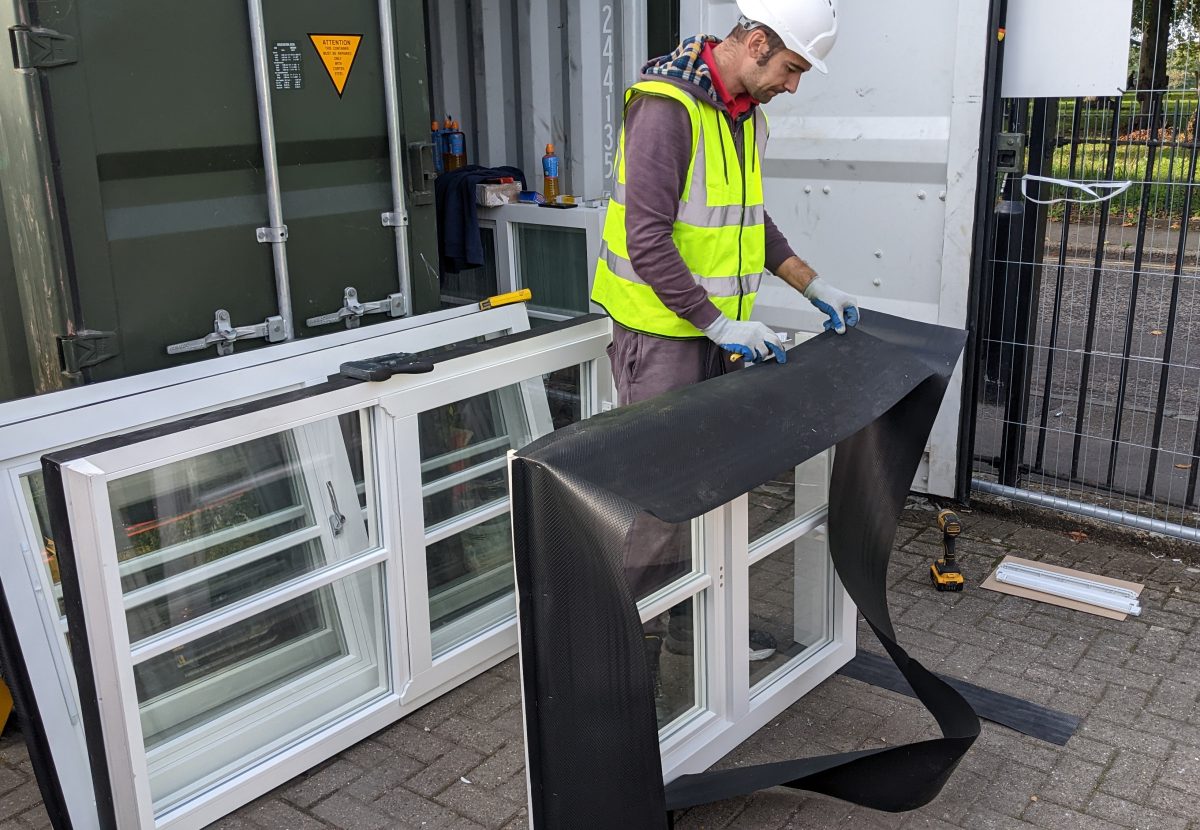 A worker in a reflective vest and helmet applies protective material to a window frame, outdoors near a storage container and gate.