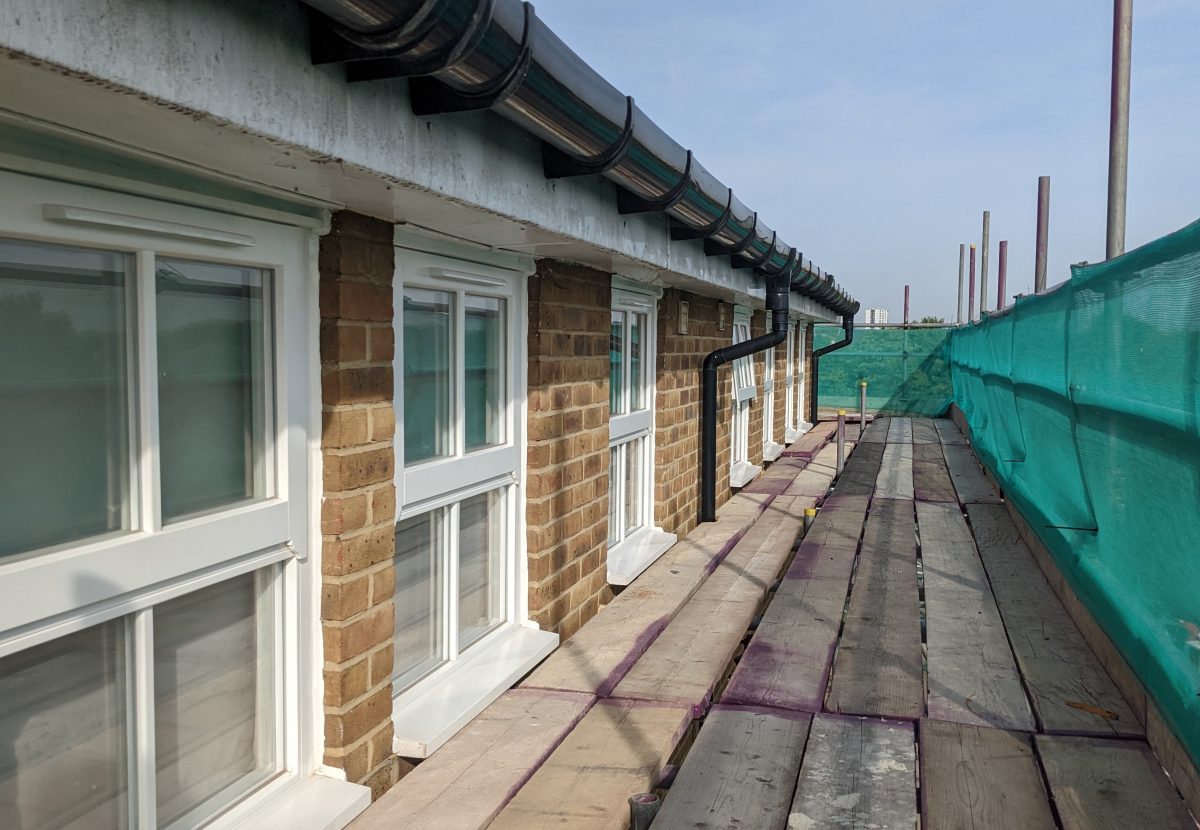 A walkway flanked by a row of windows on a brick building under tiled eaves with scaffolding and a green net stretched along the side.