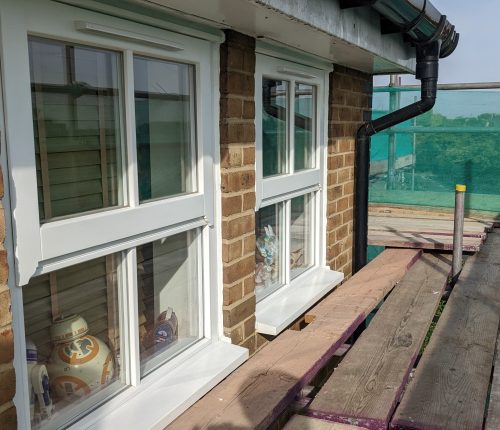 Two white-framed windows on a brick wall, each with a ceramic figurine on the sill, overlooking a wooden deck with greenery in the distance.