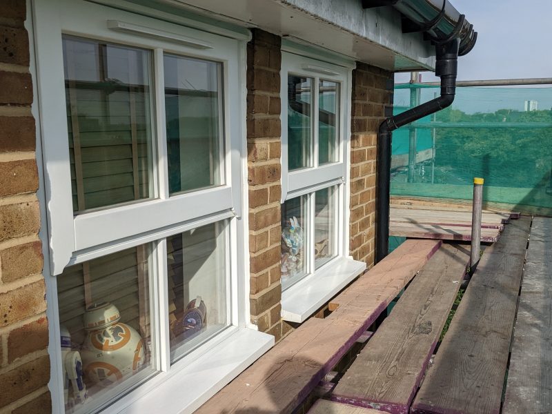 Two white-framed windows on a brick wall, each with a ceramic figurine on the sill, overlooking a wooden deck with greenery in the distance.