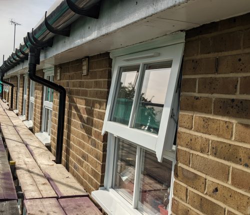 A row of terraced houses with open white windows, gutter pipes running along the top, and a clear sky visible in the reflection.