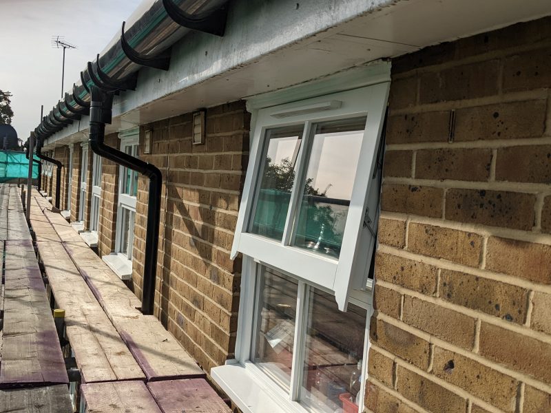 A row of terraced houses with open white windows, gutter pipes running along the top, and a clear sky visible in the reflection.