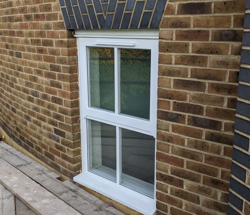 A white-framed double-hung window is set within a brick wall, under an overhanging roof with decorative brickwork