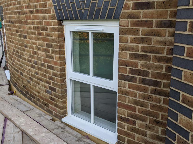 A white-framed double-hung window is set within a brick wall, under an overhanging roof with decorative brickwork