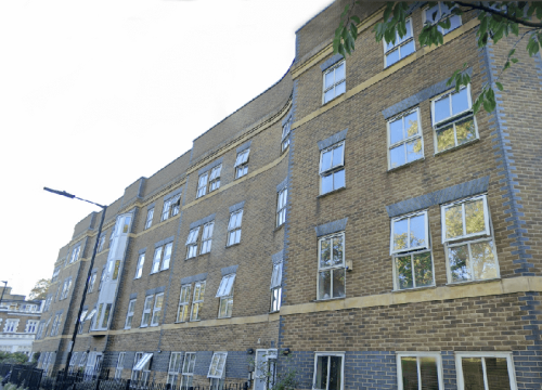 A modern brick apartment building with multiple windows, bathed in sunlight, amidst an urban setting with green trees.