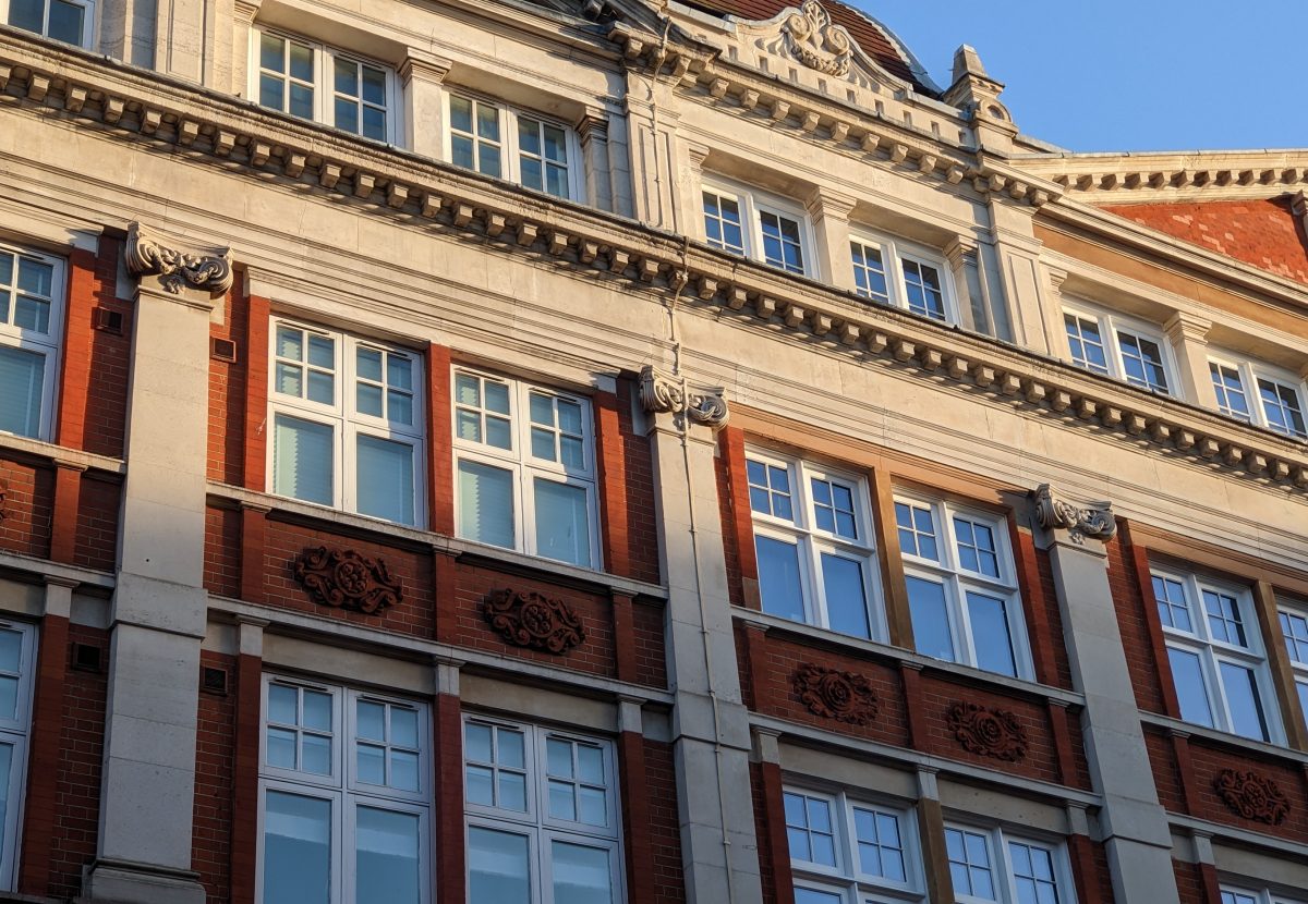 A historic red-brick building with white framed windows, ornate carvings, and a clear blue sky in the background.