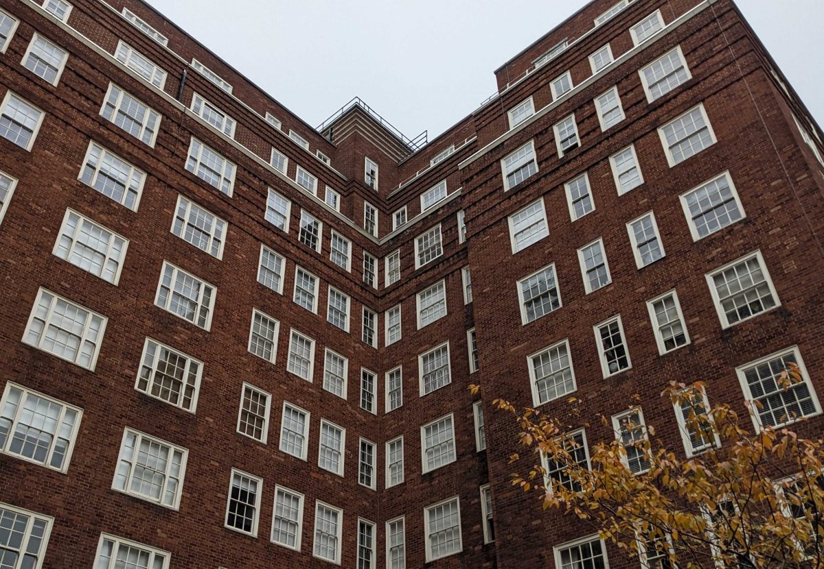 A large brick building with numerous symmetrical windows against a cloudy sky. A hint of autumn foliage is visible at the bottom.