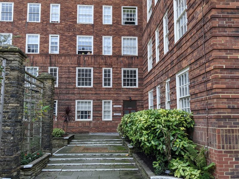 A multi-story brick building with symmetrical windows, flanked by green plants, and a pathway leading to a central door, under a cloudy sky.