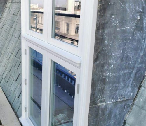 A white window provides a view of urban buildings reflecting on its glass, against an adjacent gray slate roof.