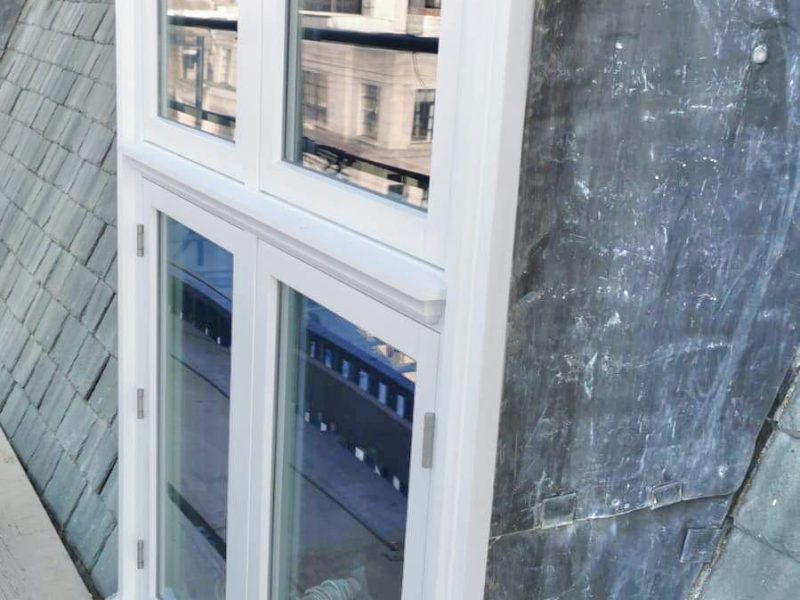 A white window provides a view of urban buildings reflecting on its glass, against an adjacent gray slate roof.