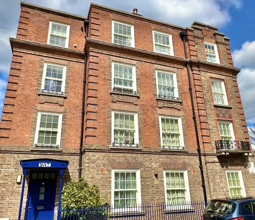 A traditional red brick building with white windows under a cloudy sky, featuring a blue doorway. There are cars parked in front, behind a blue fence.