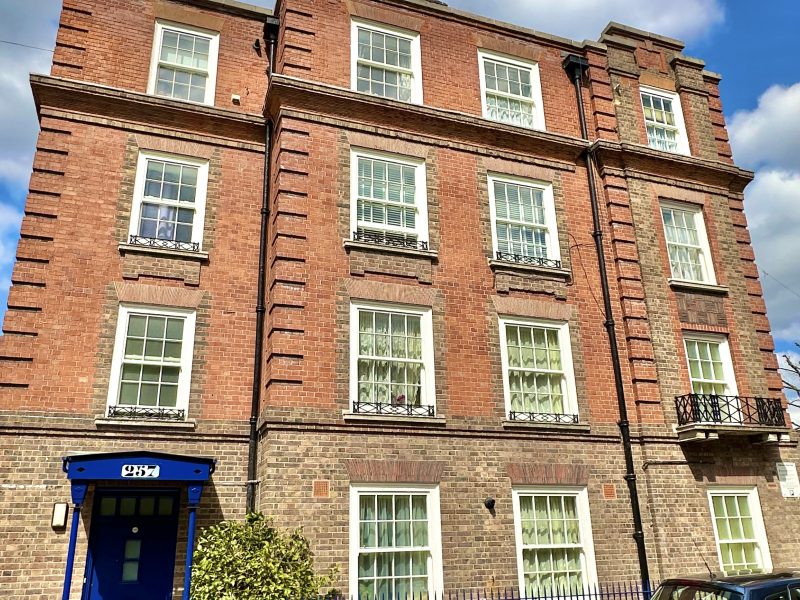 A traditional red brick building with white windows under a cloudy sky, featuring a blue doorway. There are cars parked in front, behind a blue fence.