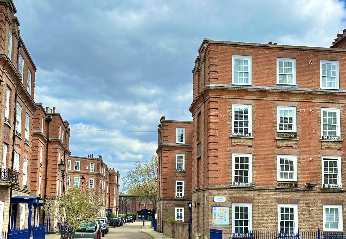 Red brick buildings flank a street under a cloudy sky, with parked cars and sparse greenery, creating an urban residential scene.