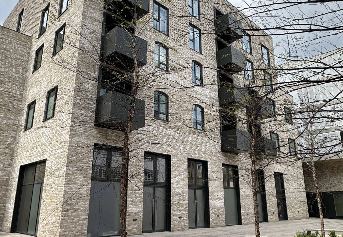 A modern brick apartment building features large windows and dark balconies, surrounded by leafless trees in a contemporary urban setting. The sky appears overcast, adding to the muted atmosphere.