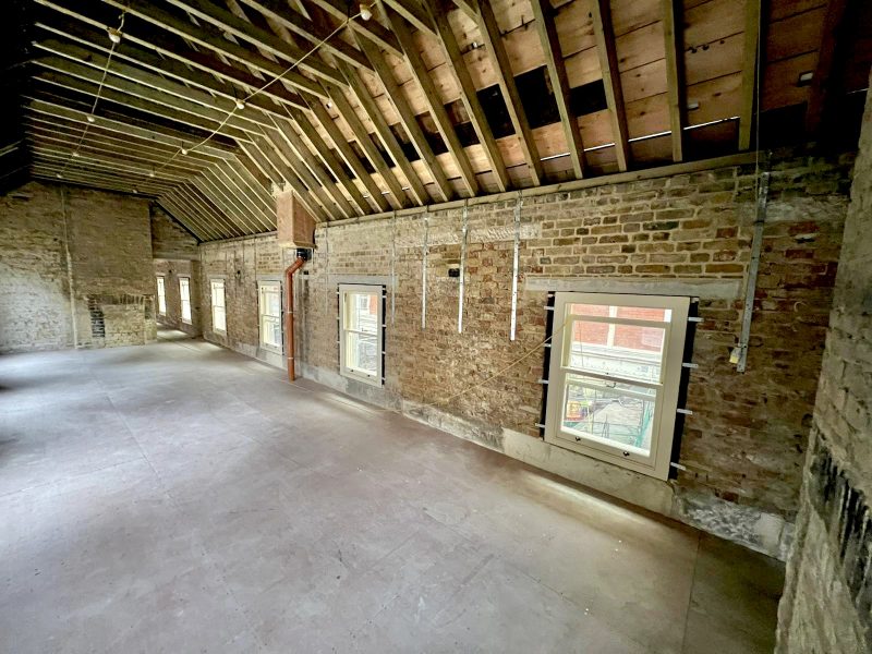 An empty, spacious room features exposed brick walls and windows, with visible rafters overhead. Natural light enters through the windows, illuminating the bare floor and construction elements.