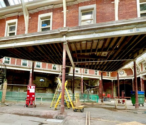 A construction site features exposed beams and columns, with a ladder and scaffolding for access. Renovation is underway in a brick building, evident from the unfinished interior and tools scattered around.