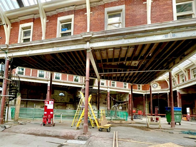 A construction site features exposed beams and columns, with a ladder and scaffolding for access. Renovation is underway in a brick building, evident from the unfinished interior and tools scattered around.