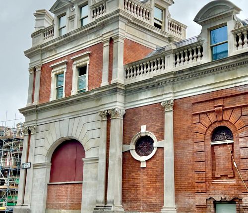 A stately brick building with arches, ornate windows, and a classical façade, partially obscured by scaffolding on the right. Cloudy sky in the background.