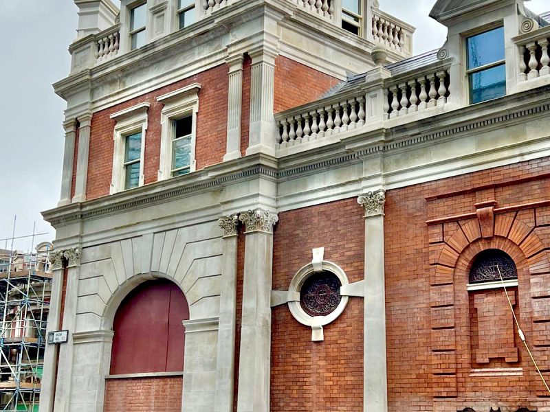 A stately brick building with arches, ornate windows, and a classical façade, partially obscured by scaffolding on the right. Cloudy sky in the background.