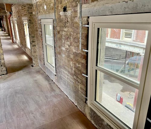 A series of white-framed windows line a brick wall in a narrow corridor, with light illuminating the space. Construction is ongoing in the area outside, visible through the windows.