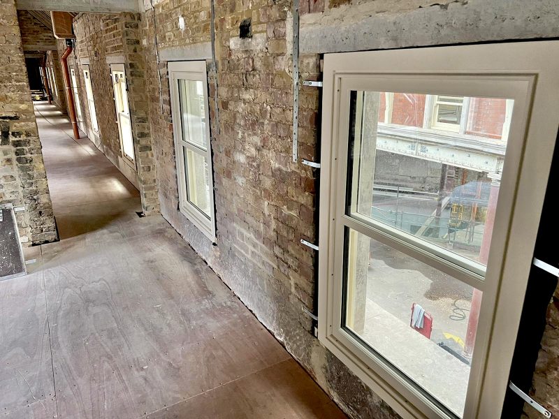A series of white-framed windows line a brick wall in a narrow corridor, with light illuminating the space. Construction is ongoing in the area outside, visible through the windows.