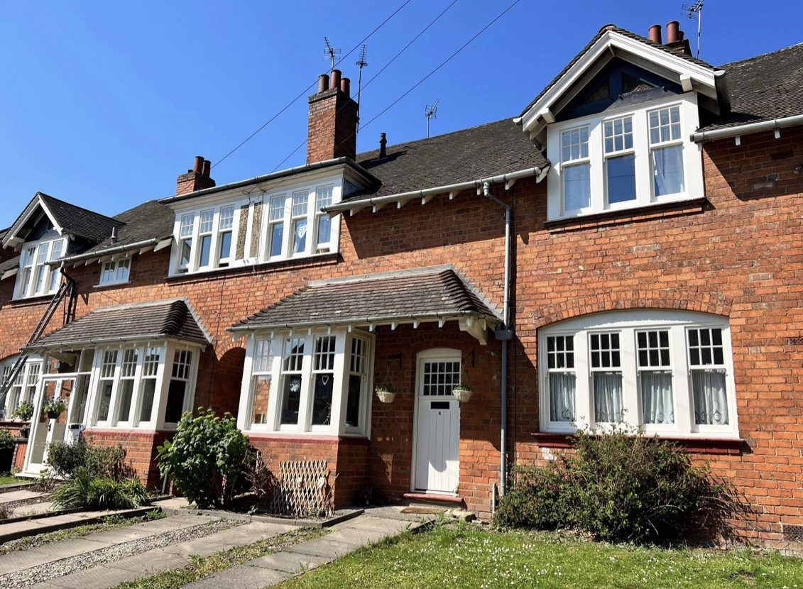 Red-brick terraced houses, with white windows and doors, stand under a clear blue sky, each featuring a small front garden with different plants.