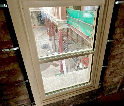 A window frames a view of an under-construction space with scaffolding and materials scattered on the ground, surrounded by bare brick walls and protective green covering above.