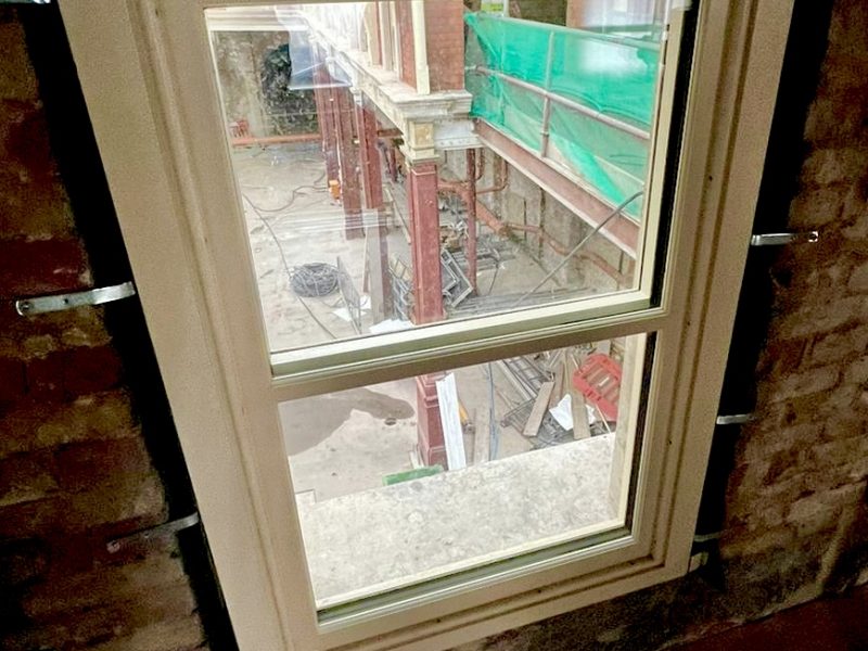 A window frames a view of an under-construction space with scaffolding and materials scattered on the ground, surrounded by bare brick walls and protective green covering above.