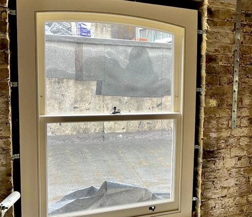 A white-framed window opens to a gray, textured wall outside. The action indicates a view into an unfinished space, with construction materials and debris visible below.