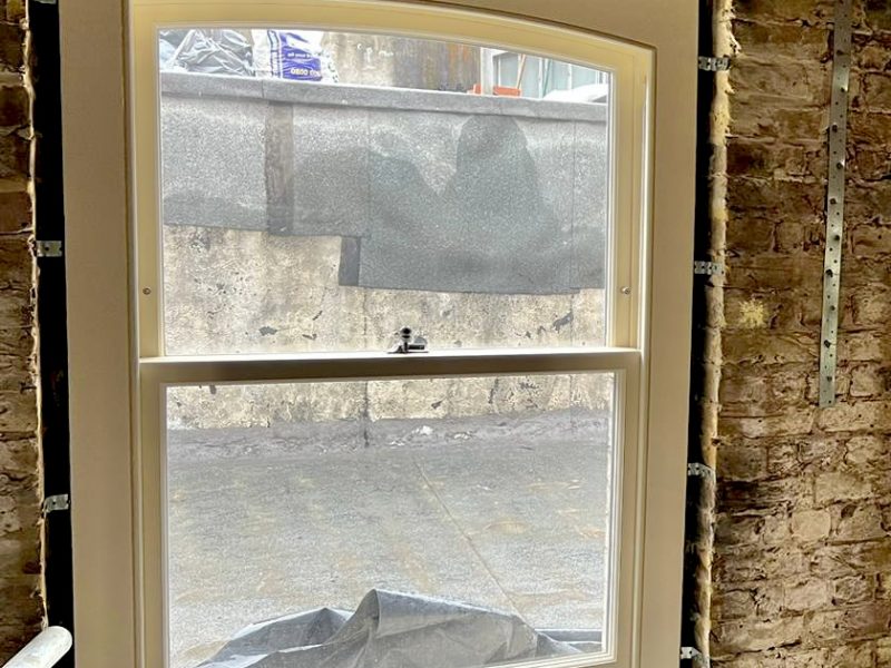 A white-framed window opens to a gray, textured wall outside. The action indicates a view into an unfinished space, with construction materials and debris visible below.