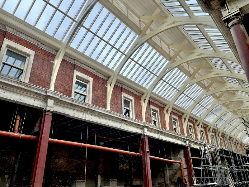 A Victorian-style brick building undergoing renovation, with visible scaffolding and a glass roof supported by arched white beams.