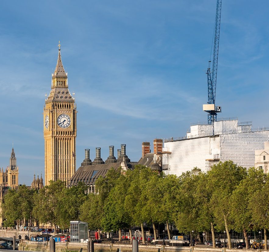 Big Ben stands prominently against a clear sky, with surrounding buildings partly obscured by scaffolding, amidst an urban riverside setting with lush trees.