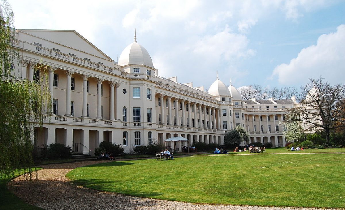 An elegant beige building with white domes, surrounded by green lawns and trees, under a partly cloudy sky. People are relaxing and socialising in the grassy area.