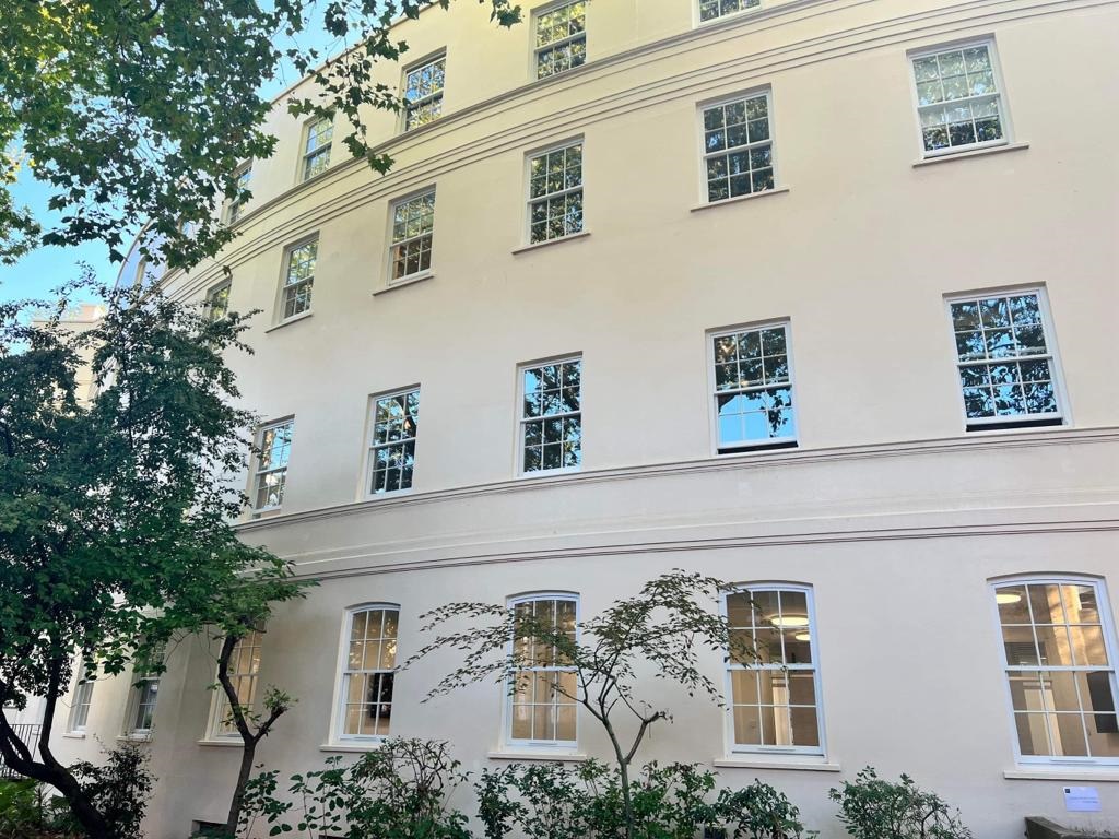 A cream-colored building with multiple windows reflecting trees, flanked by green foliage in daylight.