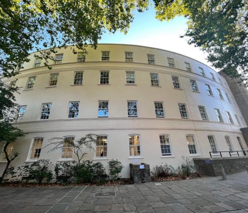 A curved, multi-story building with numerous windows, surrounded by trees, set against a clear sky.