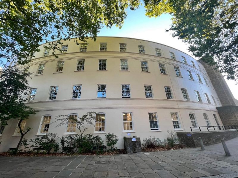 A curved, multi-story building with numerous windows, surrounded by trees, set against a clear sky.