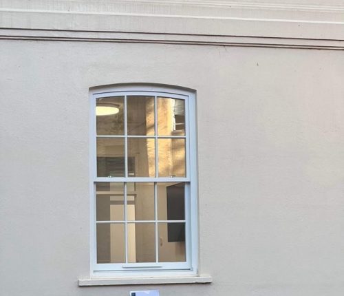 Arched window with white frame on a light grey wall, showing reflections of a building and blue sky, with interior lights visible inside.