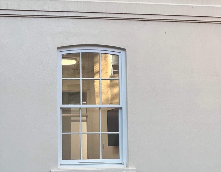 Arched window with white frame on a light grey wall, showing reflections of a building and blue sky, with interior lights visible inside.