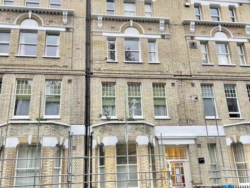 A symmetrical brick building with large windows and balconies, surrounded by a metal fence; overcast sky in the background.